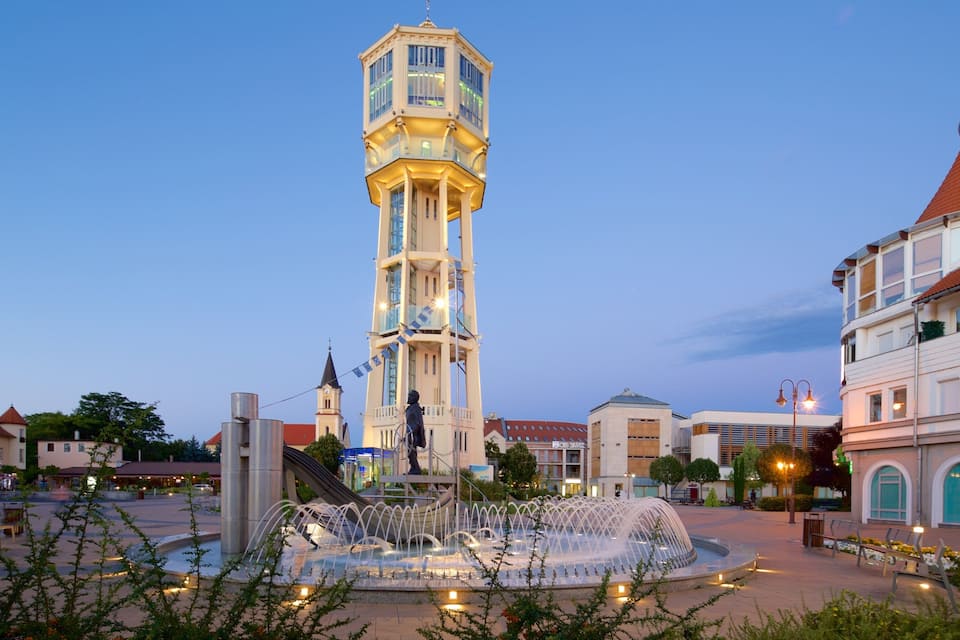 A tower with a statue in front of it, surrounded by buildings and a fountain.