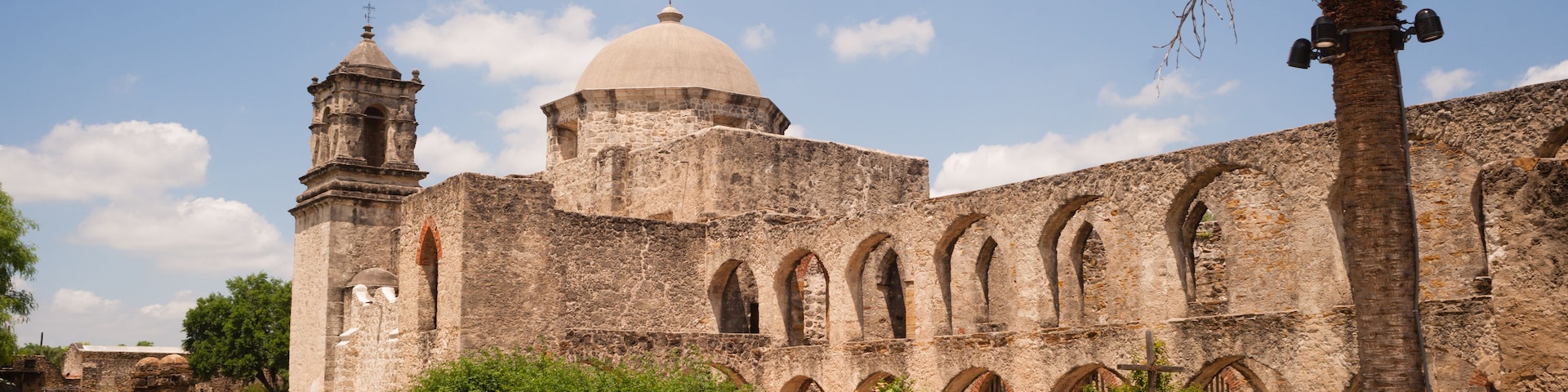 An ancient stone building with a dome and bell tower, surrounded by greenery and palm trees.