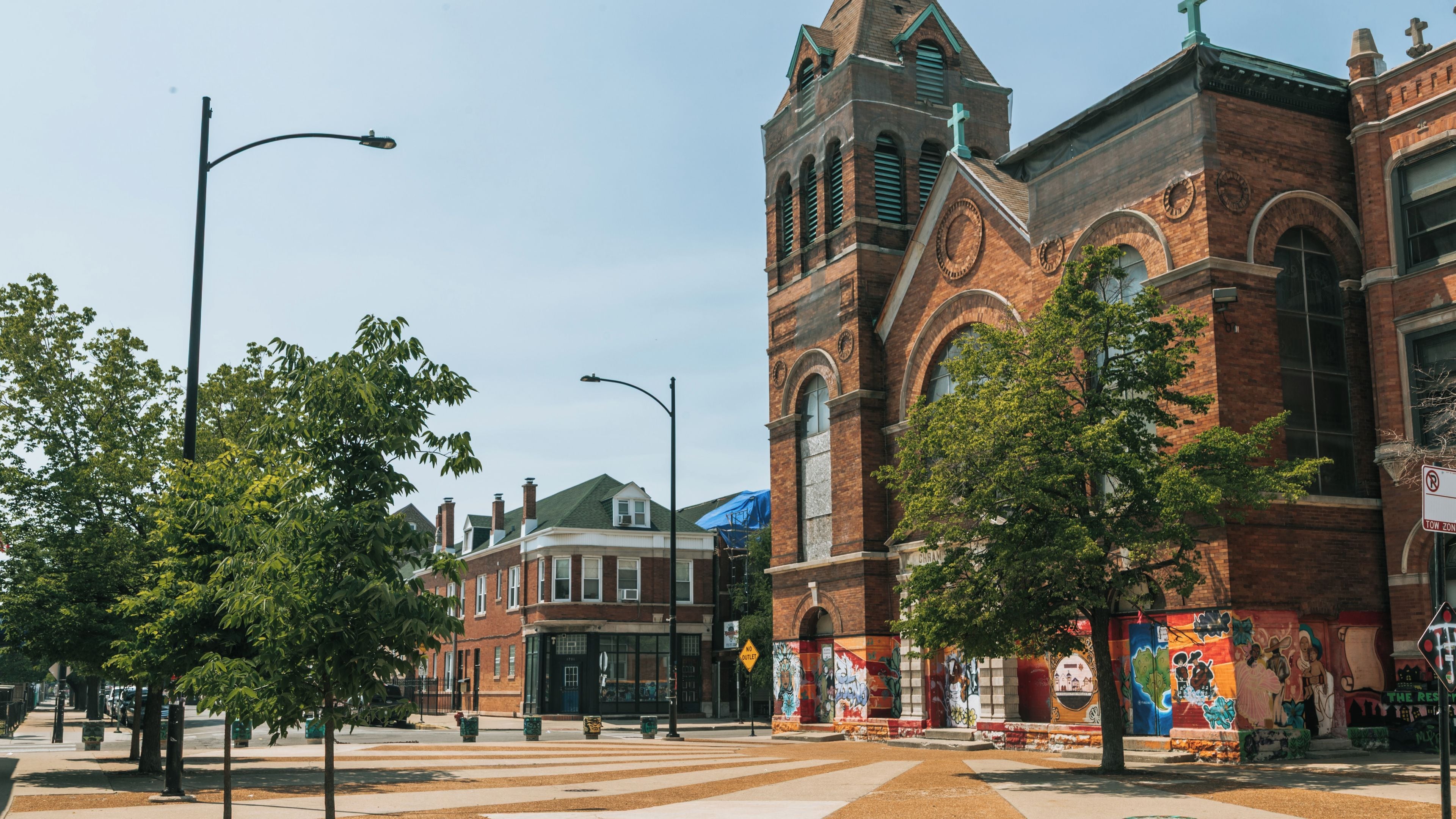 A historic brick building with a clock tower, a cross, and a street with trees and a crosswalk.