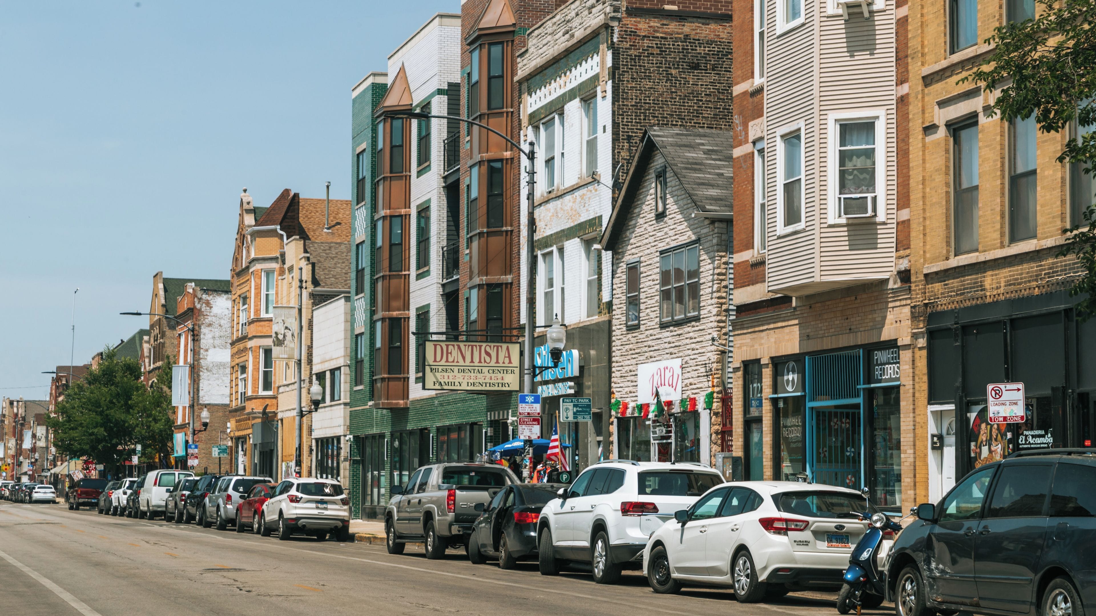 A street lined with multi-story buildings, parked cars, and a clear blue sky.