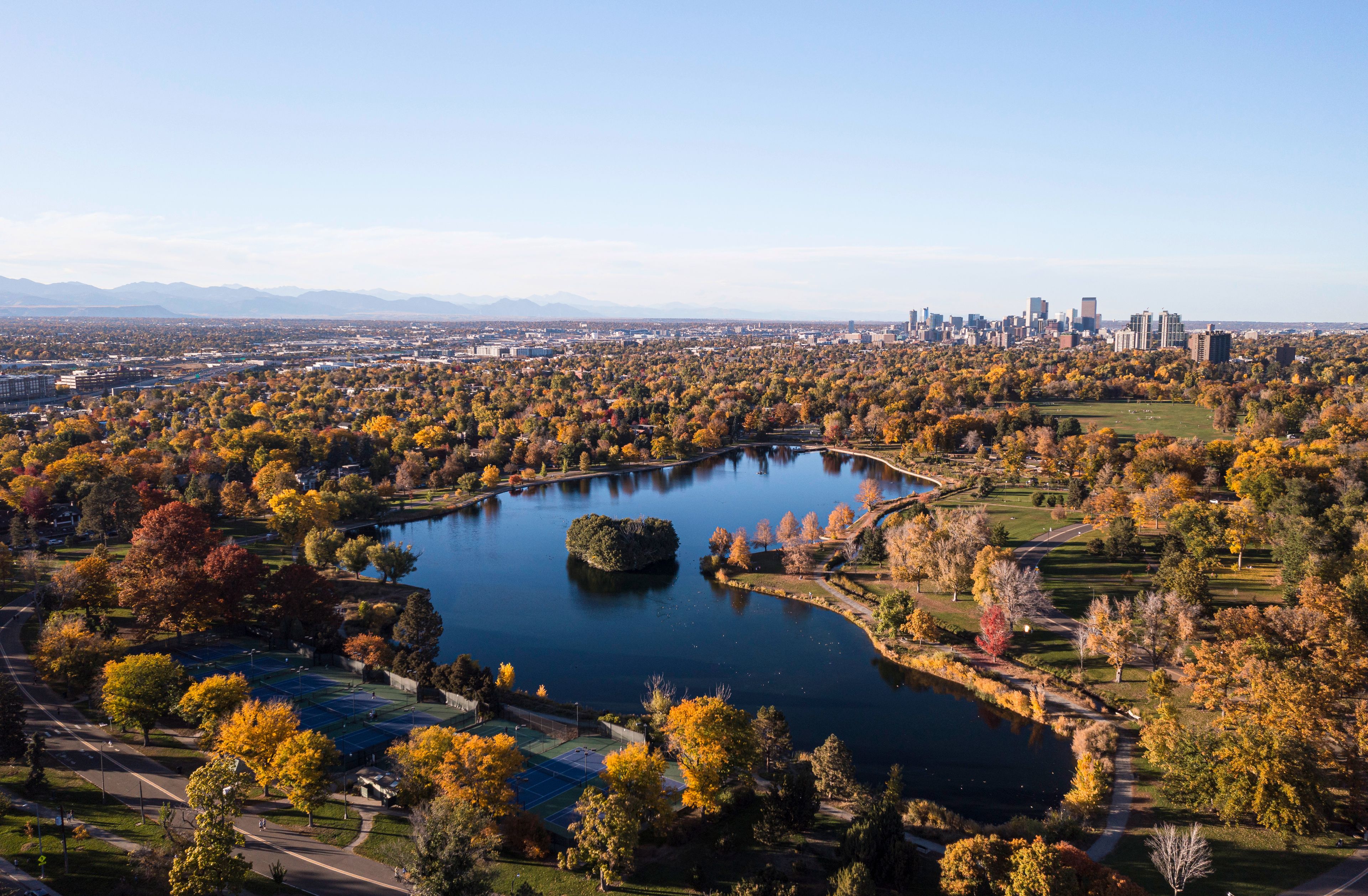 An aerial view of a city with a large lake surrounded by autumn-colored trees, a park, and a distant city skyline.