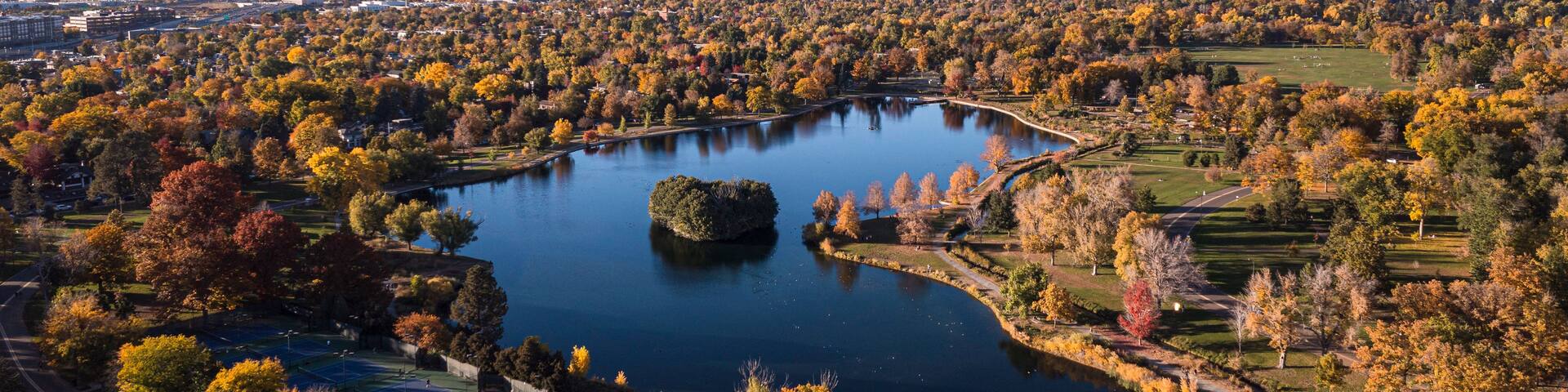 An aerial view of a city with a large lake surrounded by autumn-colored trees, a park, and a distant city skyline.