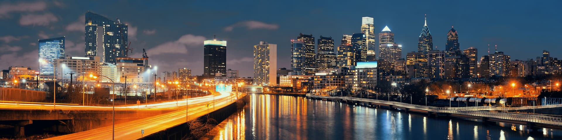 A city skyline at night with illuminated buildings, a river reflecting lights, and a bridge with light trails.