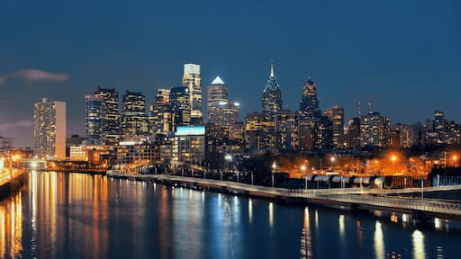 A city skyline at night with illuminated buildings, a river reflecting lights, and a bridge with light trails.