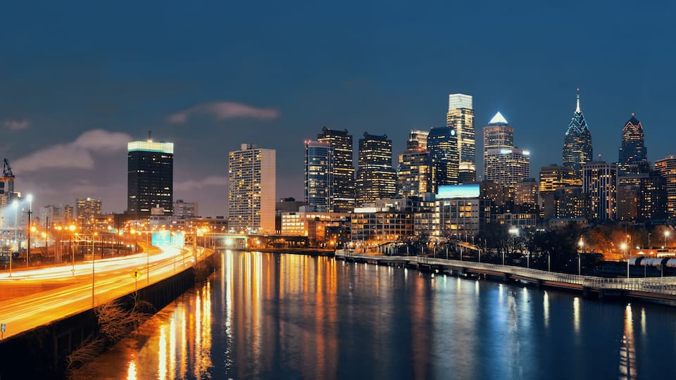A city skyline at night with illuminated buildings, a river reflecting lights, and a bridge with light trails.