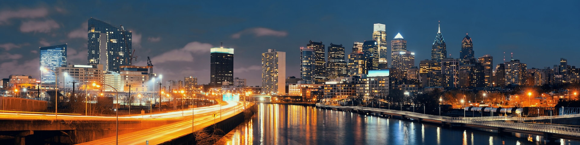 A city skyline at night with illuminated buildings, a river reflecting lights, and a bridge with light trails.