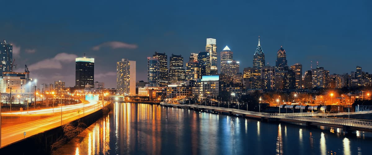 A city skyline at night with illuminated buildings, a river reflecting lights, and a bridge with light trails.