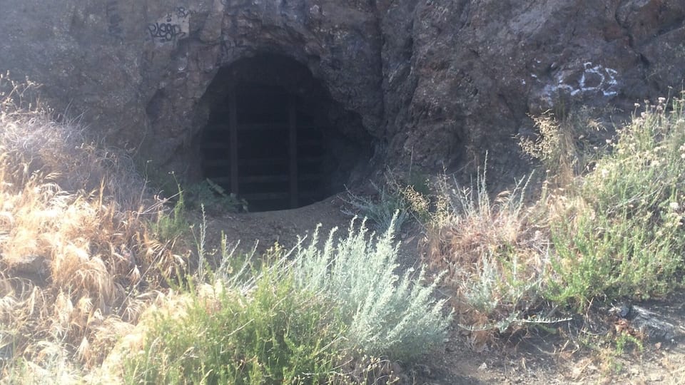 A rocky cave entrance with graffiti on the walls and dry grass in the foreground.