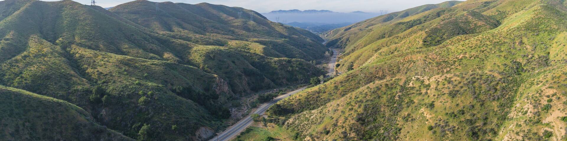 A valley with a road cutting through hilly terrain, surrounded by lush greenery.