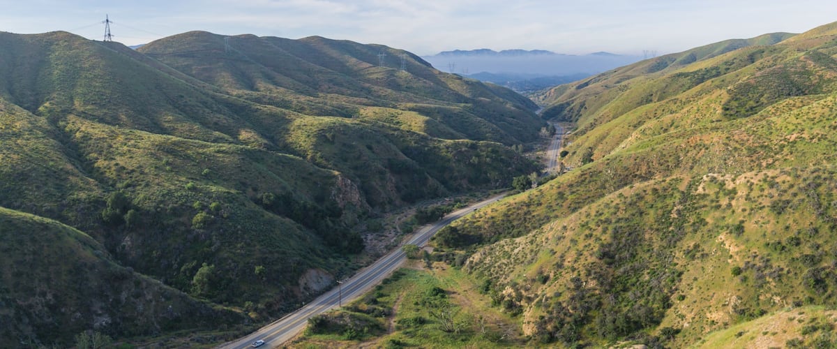 A valley with a road cutting through hilly terrain, surrounded by lush greenery.