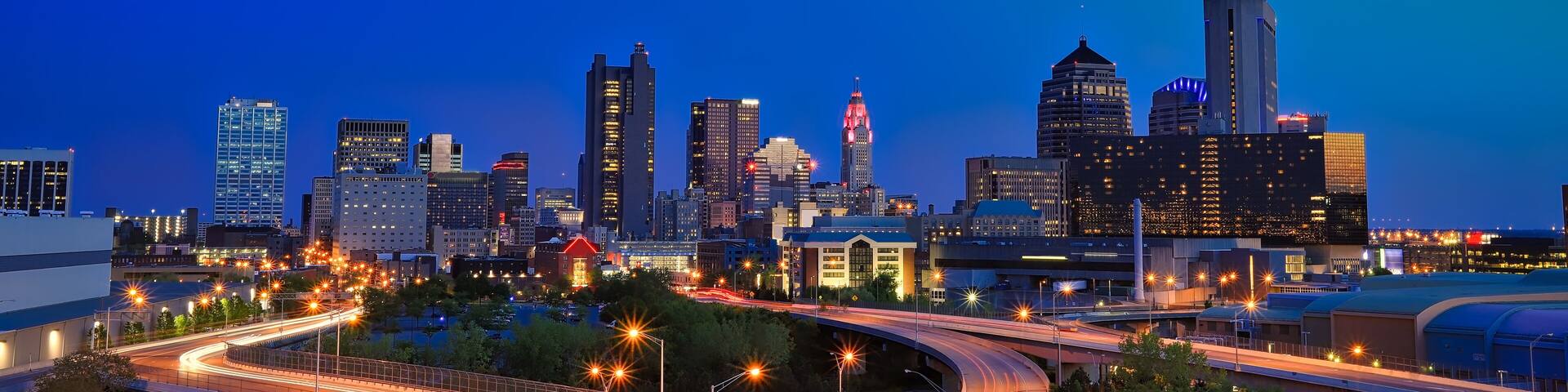 A city skyline at dusk with illuminated buildings and a curved highway.