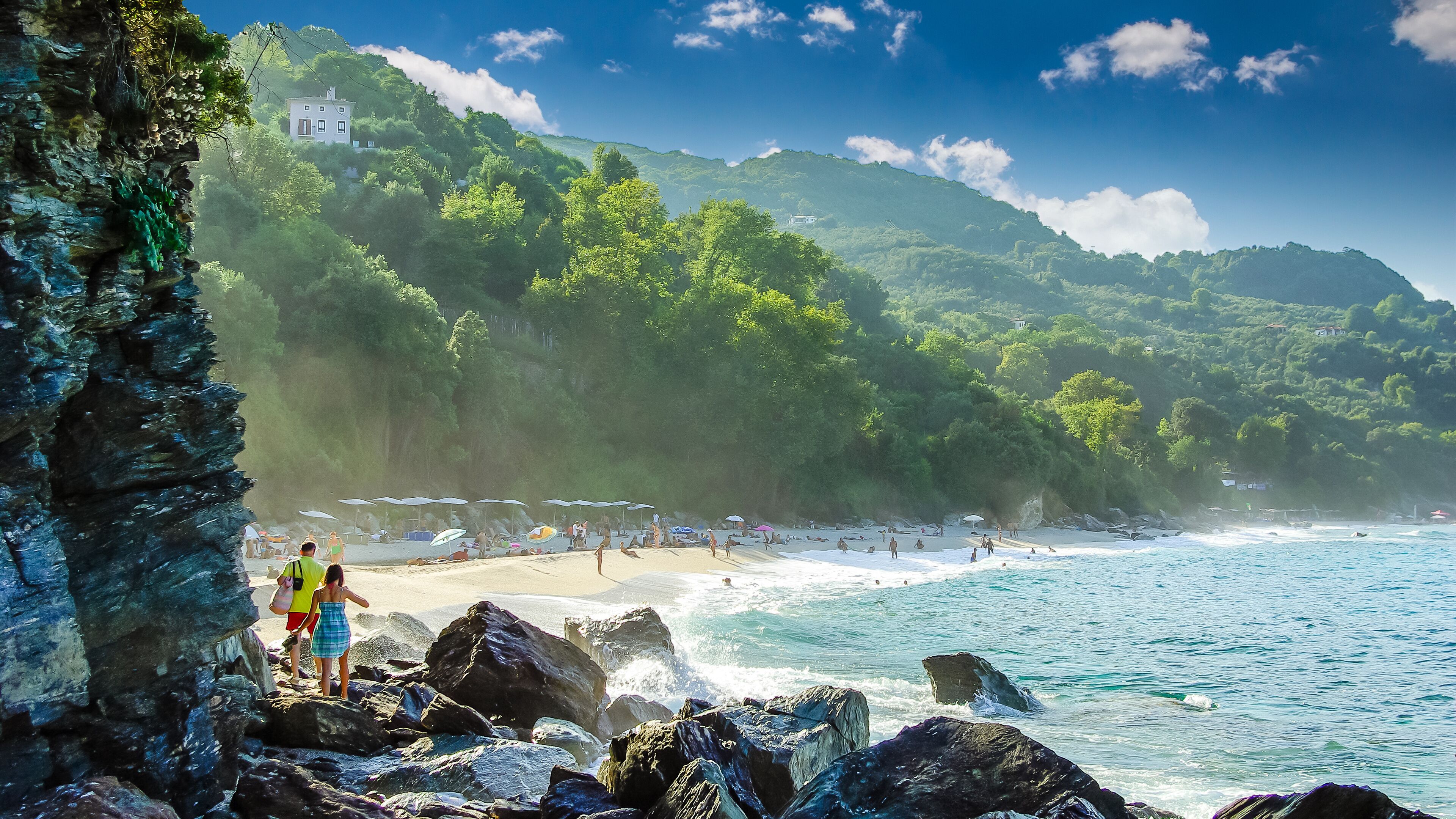 A beach with rocky shores, clear blue water, and lush green hills in the background.