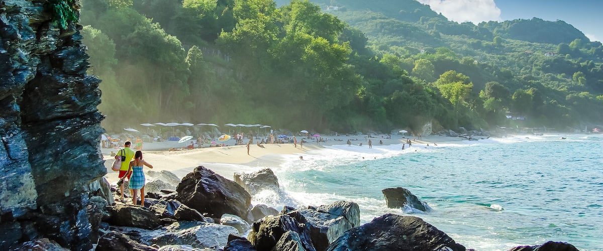 A beach with rocky shores, clear blue water, and lush green hills in the background.