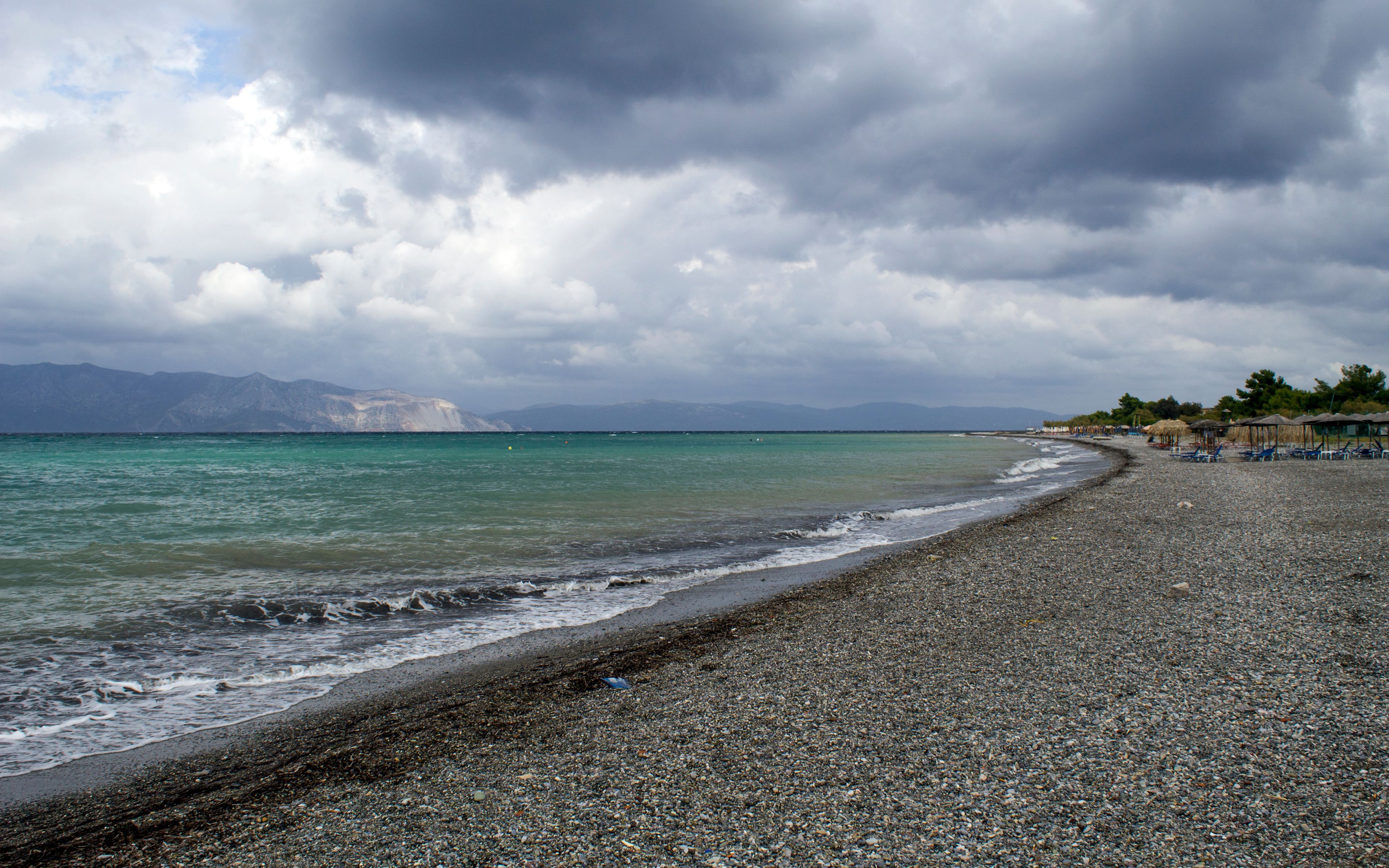 A pebbled beach with a calm sea, distant mountains, and overcast skies.