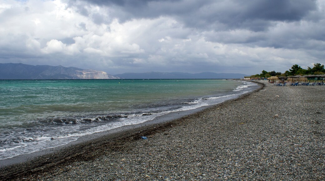 Ein Kiesstrand mit ruhiger See, fernen Bergen und bewölktem Himmel.
