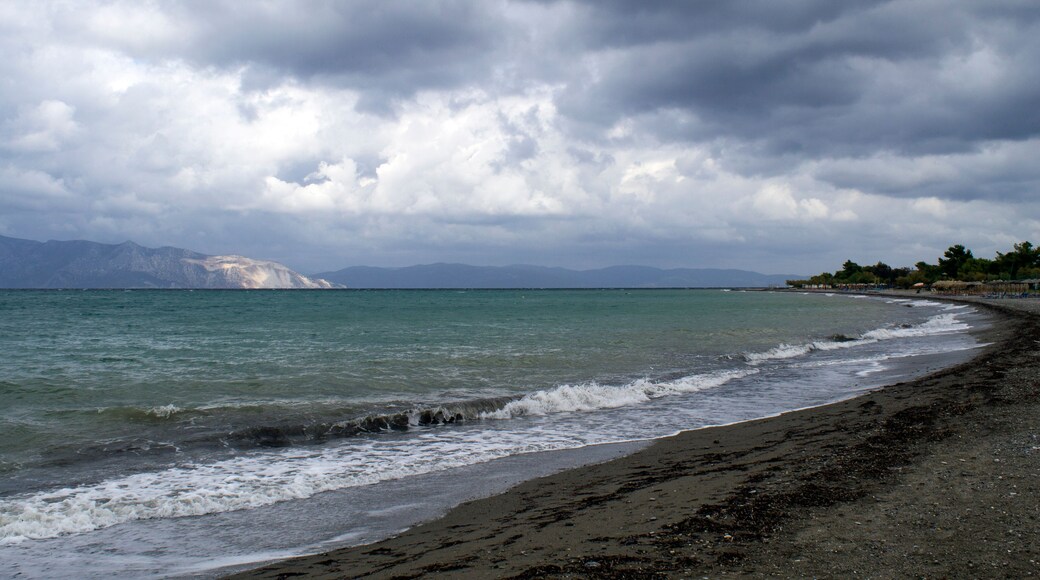 Ein Strand mit Wellen, bewölktem Himmel und Bergen im Hintergrund.