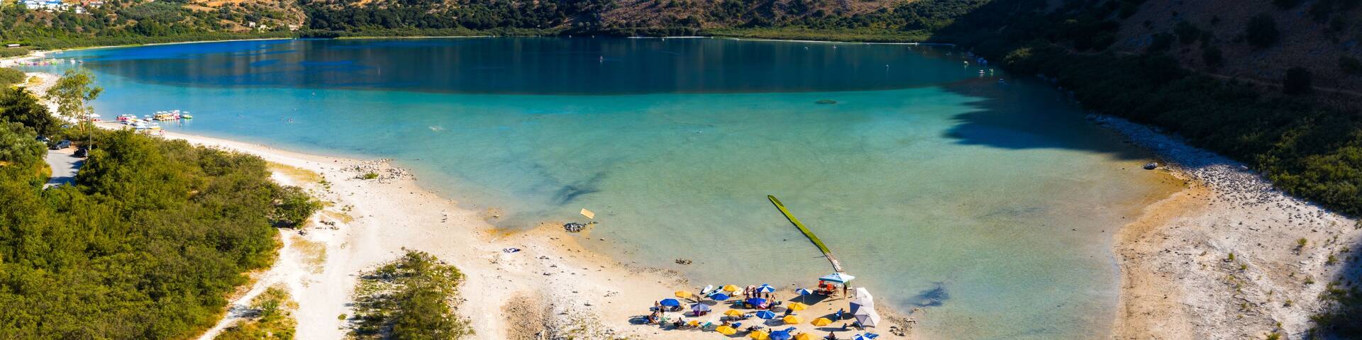 Turquoise waters of Lake Kournas in Crete, Greece, surrounded by hills. Sandy beach with umbrellas, sun loungers, and paddle boats along the shore.