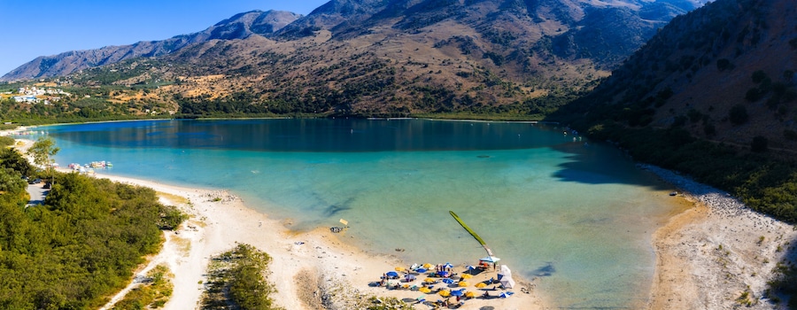 Ein See, umgeben von Bergen, ein Strand mit Sonnenschirmen und Booten.