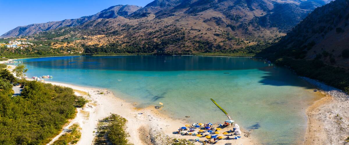 A lake surrounded by mountains, a beach with umbrellas, and boats.