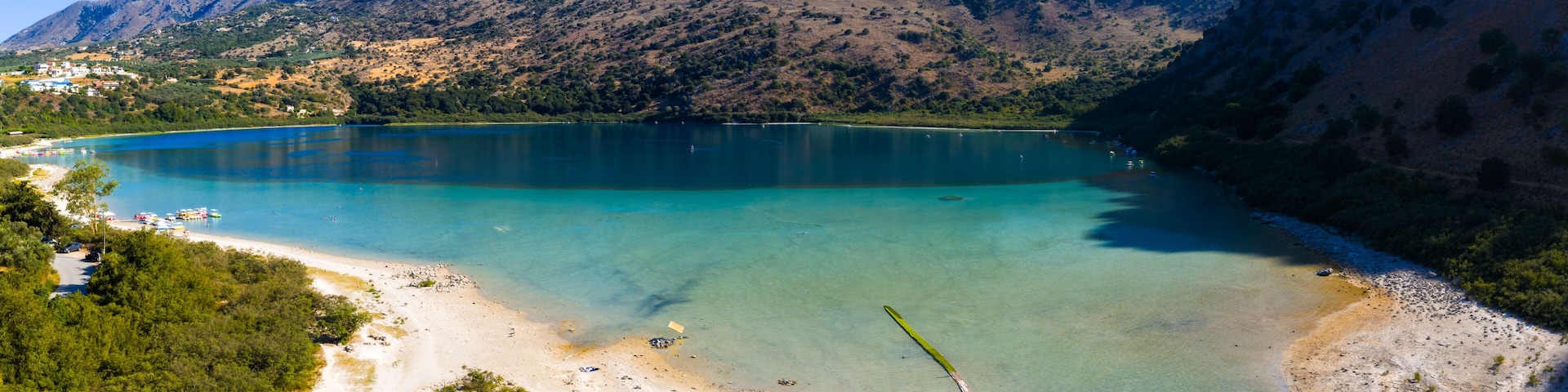 A lake surrounded by mountains, a beach with umbrellas, and boats.