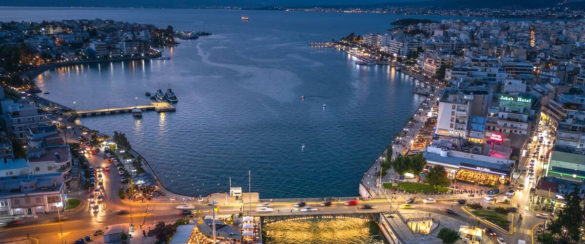 A coastal cityscape at dusk with a prominent waterfront, a large boat docked near the center, and a mix of residential and commercial buildings along the shore.