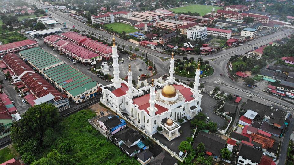 An aerial view of a city with a prominent white mosque with a golden dome and minarets, surrounded by residential and commercial buildings.