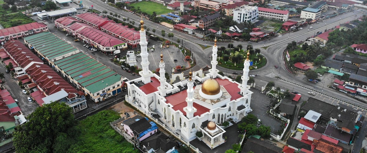 An aerial view of a city with a prominent white mosque with a golden dome and minarets, surrounded by residential and commercial buildings.