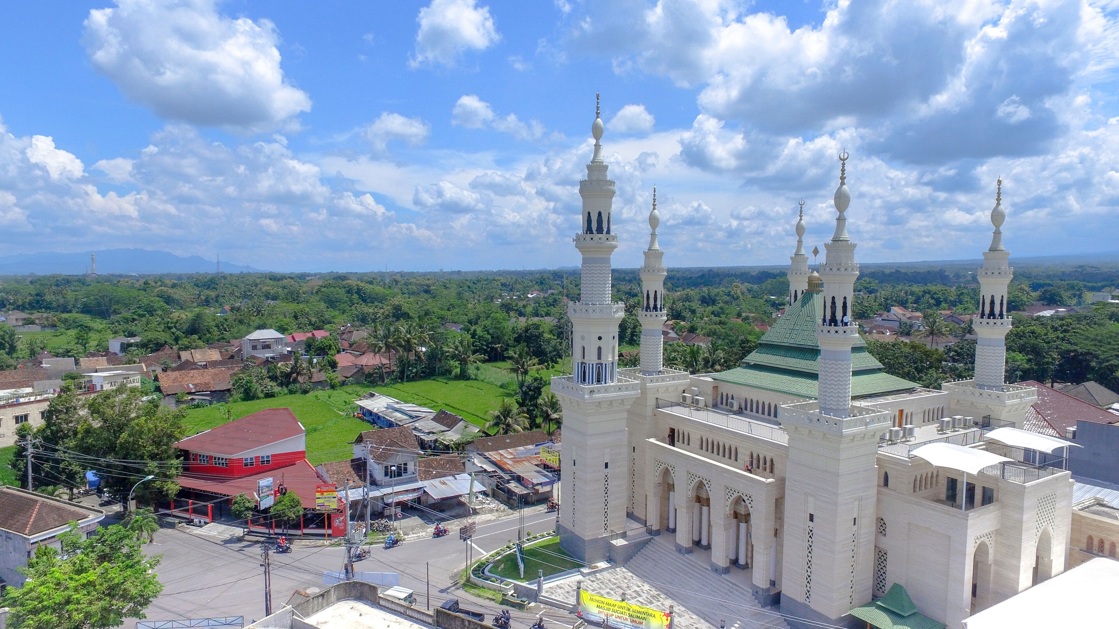 A mosque with multiple minarets and a green roof, surrounded by lush greenery and a small town.