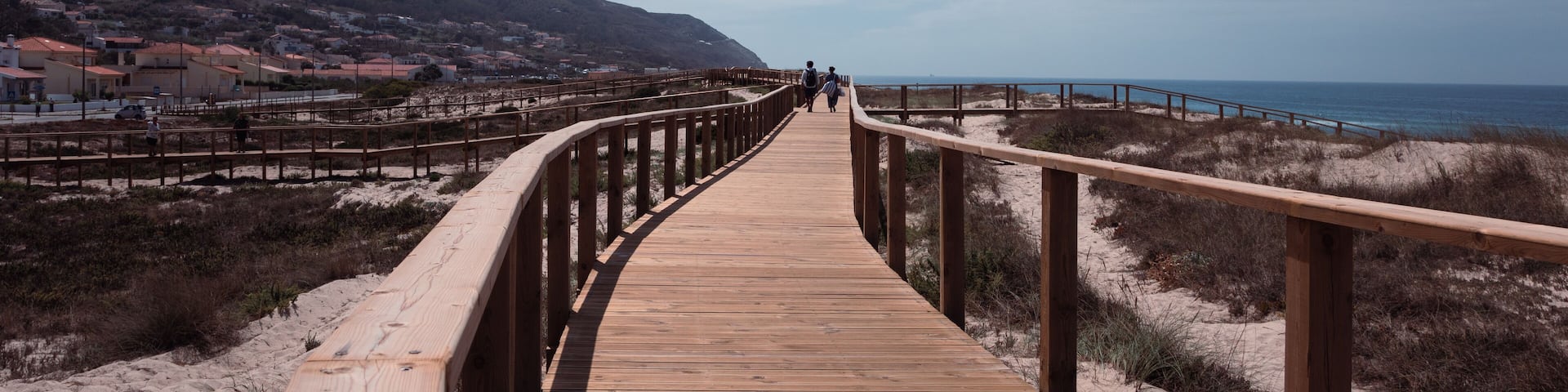 A great spot for a pleasant walk by the sea on a warm summer afternoon. Long wooden pier boardwalk and handrail in Quiaios beach with distant houses, mountain and the blue sky above