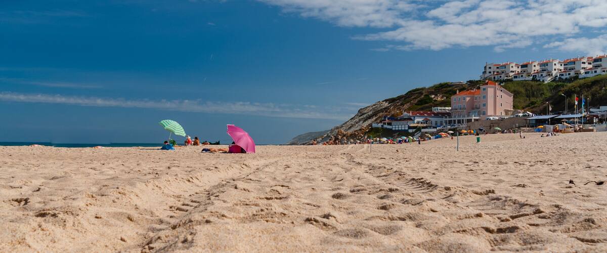 A sandy beach with footprints, a pink kite, and a building on a hill in the background.