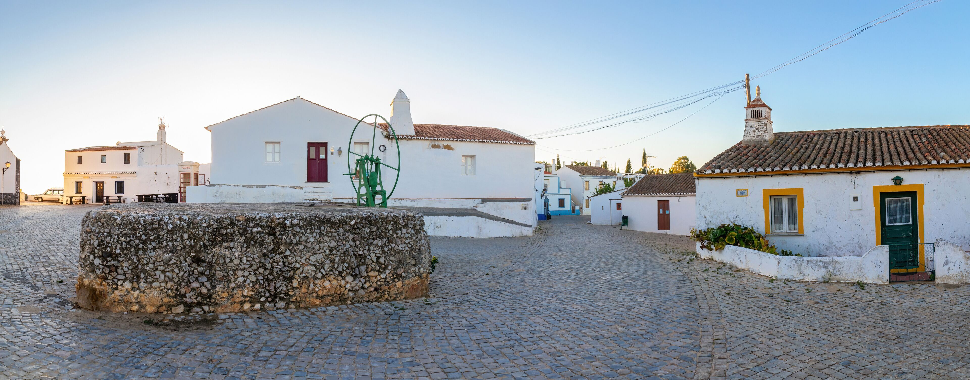 A cobblestone street with white buildings, a stone wall, and a green water pump.