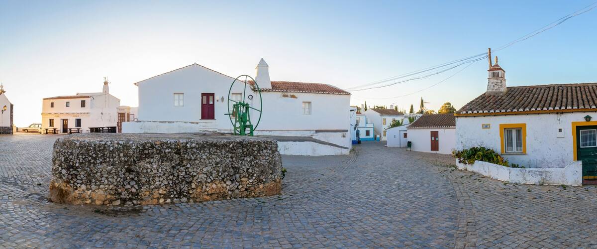 A cobblestone street with white buildings, a stone wall, and a green water pump.