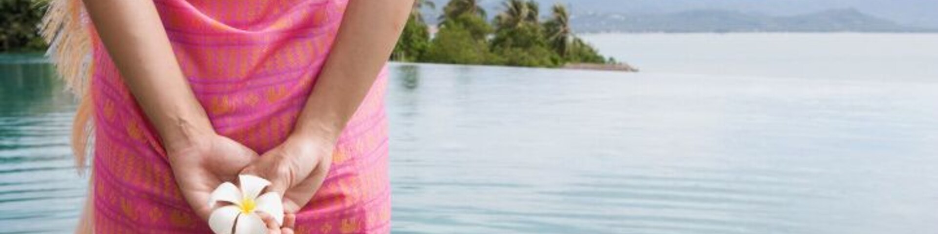 Una persona con un vestido rosa estampado sostiene una flor blanca junto a un cuerpo de agua tranquilo, con una isla y montañas a lo lejos.