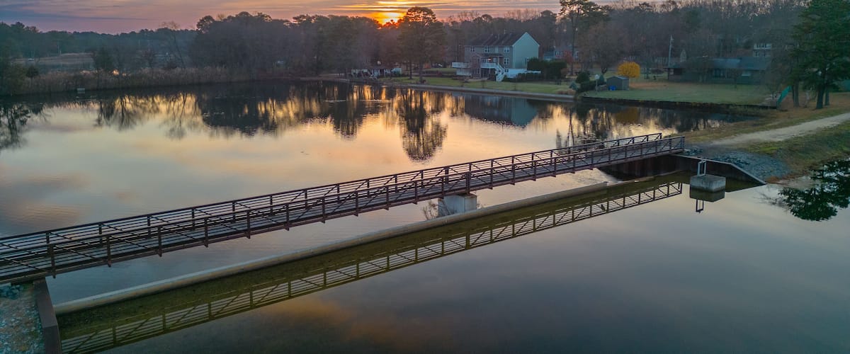 A calm lake with a bridge, trees, and houses in the background.