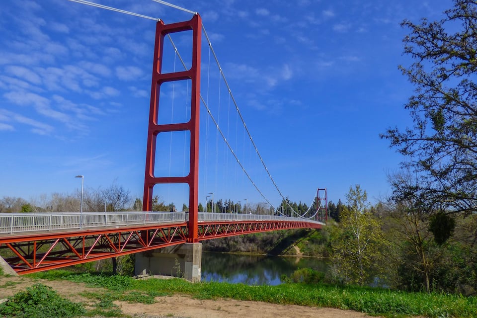 Un ponte sospeso rosso che scavalca un fiume, con verdeggianti alberi sullo sfondo.