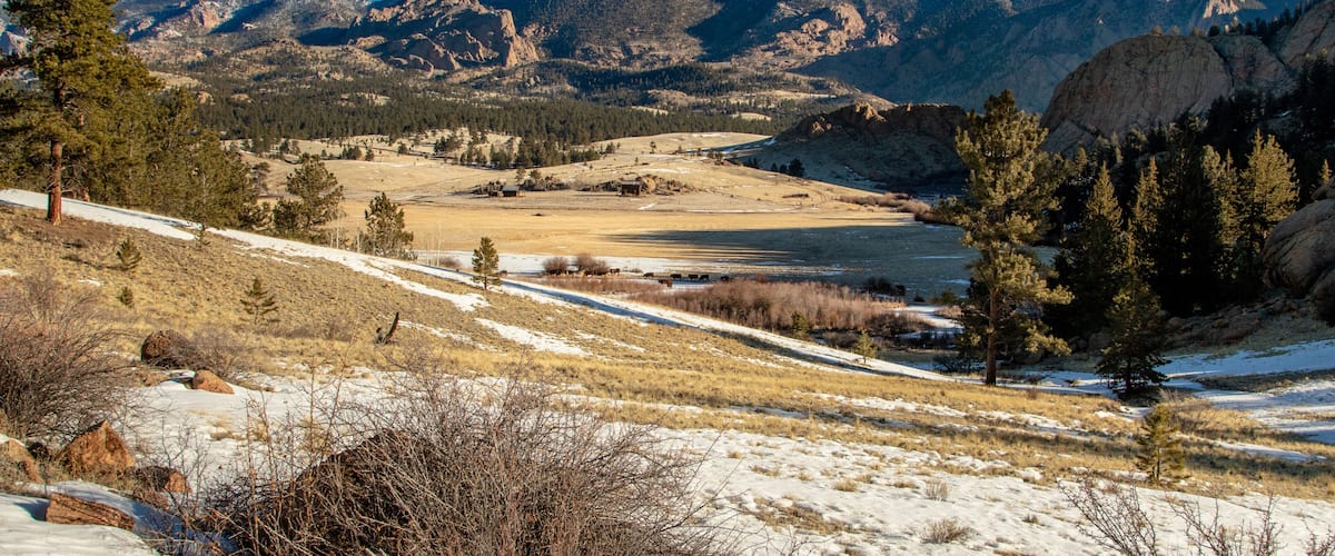 A mountainous landscape with a valley, snow-covered ground, and sparse vegetation.