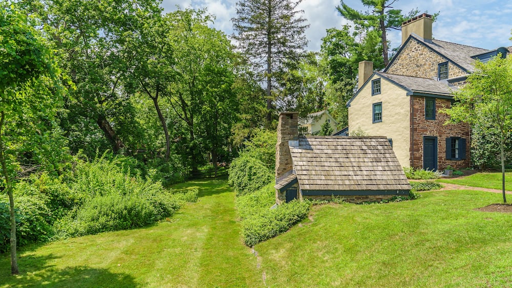 Une maison avec une cheminée en pierre, entourée d’une végétation luxuriante et de grands arbres.
