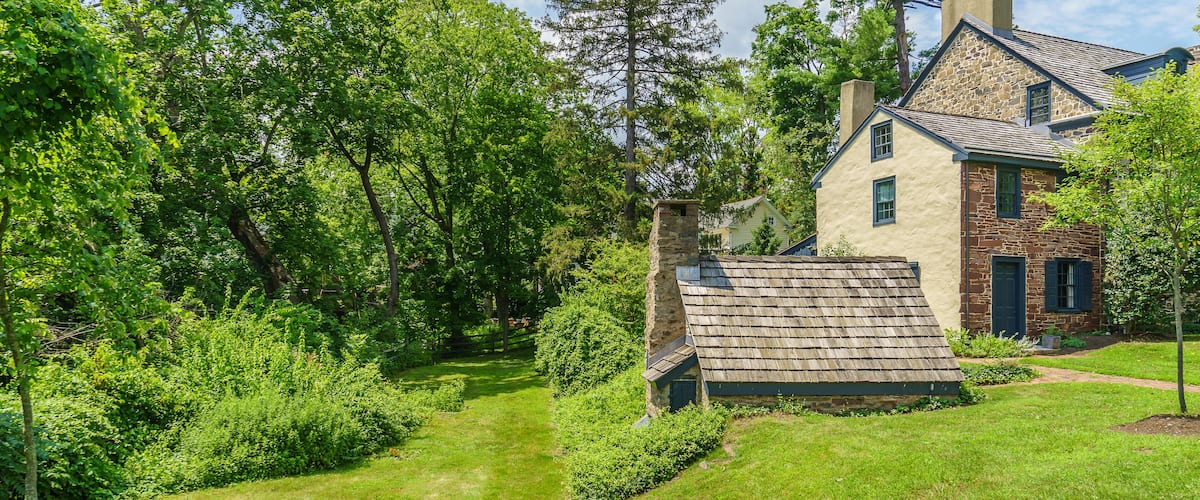 A house with a stone chimney, surrounded by lush greenery and tall trees.