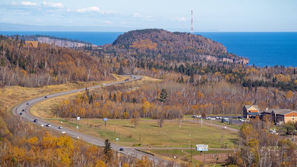 A winding road through a forested area with autumn foliage, a distant mountain, and a body of water.