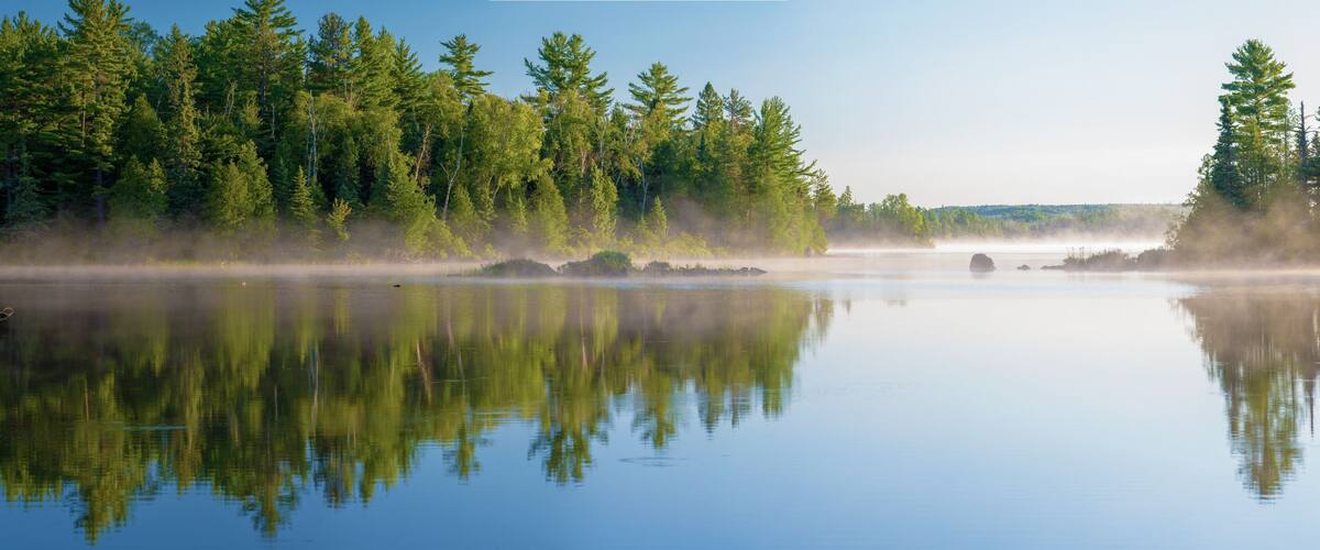 A calm lake with dense forest on both sides, reflecting the trees and mist rising from the water.