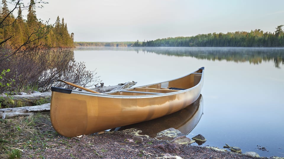 A canoe on a rocky shore by a calm lake with trees in the background.