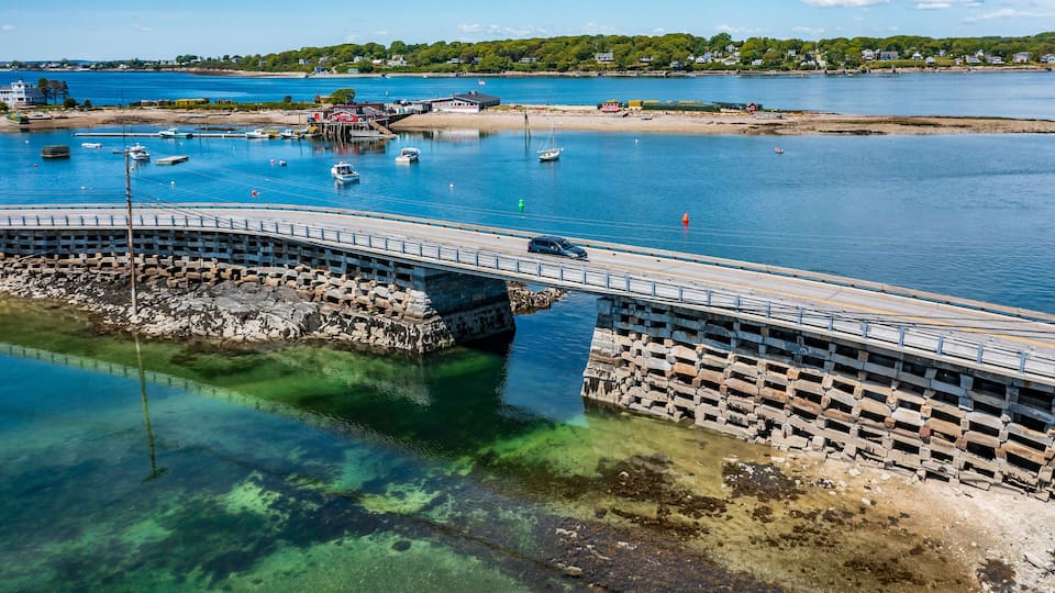 A coastal bridge over clear water with boats, a sandy beach, and houses in the background.