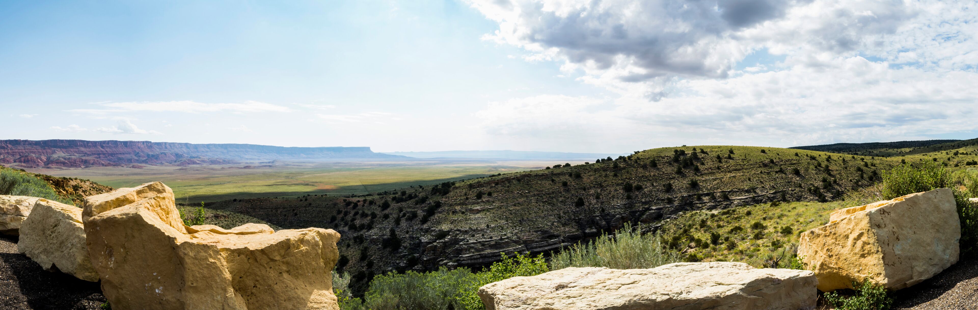 A vast canyon landscape with prominent rock formations in the foreground and a distant view of rolling hills under a partly cloudy sky.
