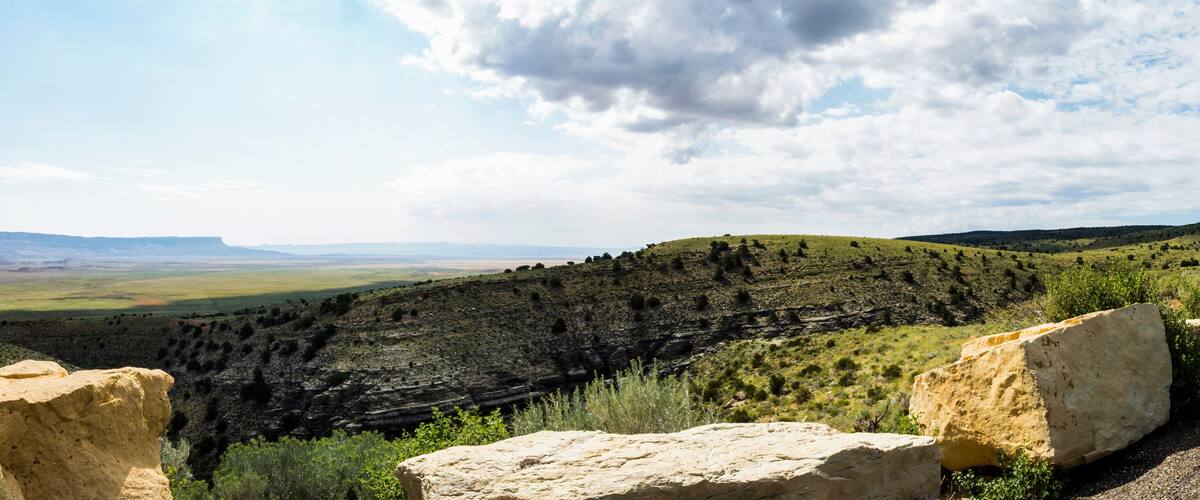 A vast canyon landscape with prominent rock formations in the foreground and a distant view of rolling hills under a partly cloudy sky.