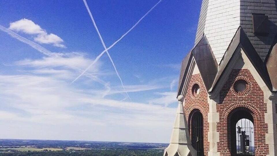 A church with a tall steeple and brick architecture, surrounded by a lush green landscape and a clear blue sky.