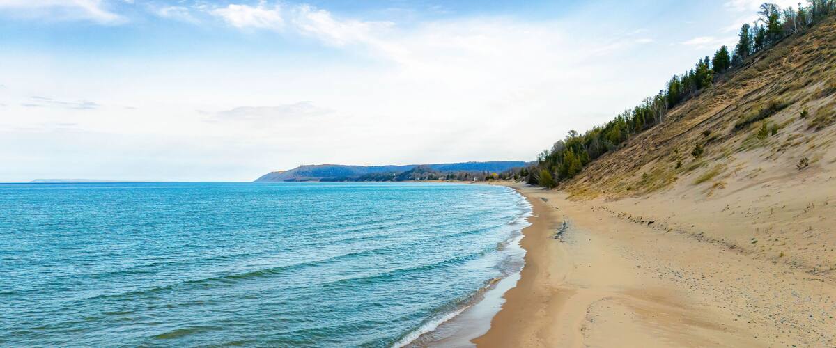 A sandy beach with gentle waves, a clear sky, and a forested hillside in the background.