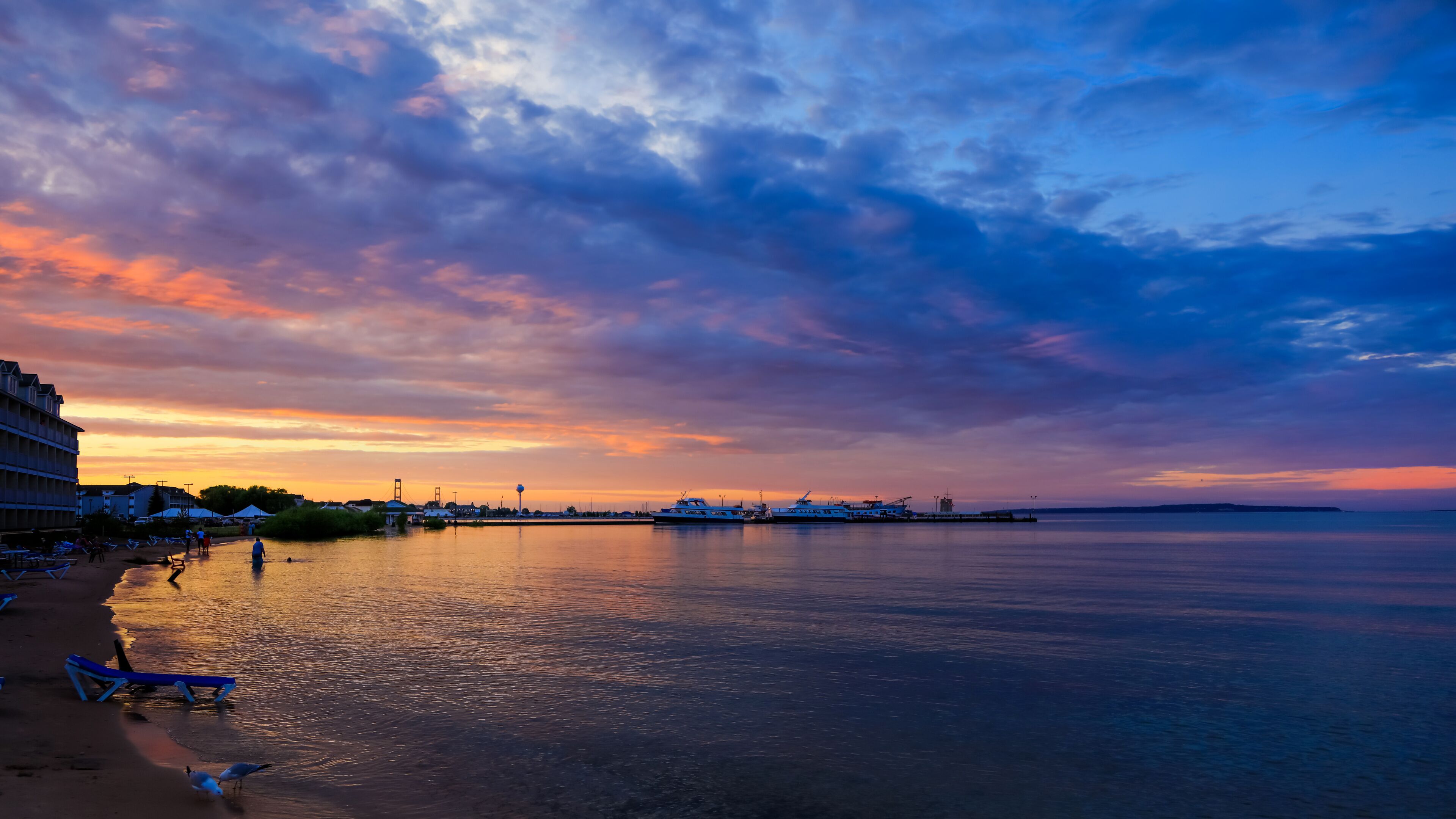 A waterfront at sunset with boats, a pier, and beach chairs.