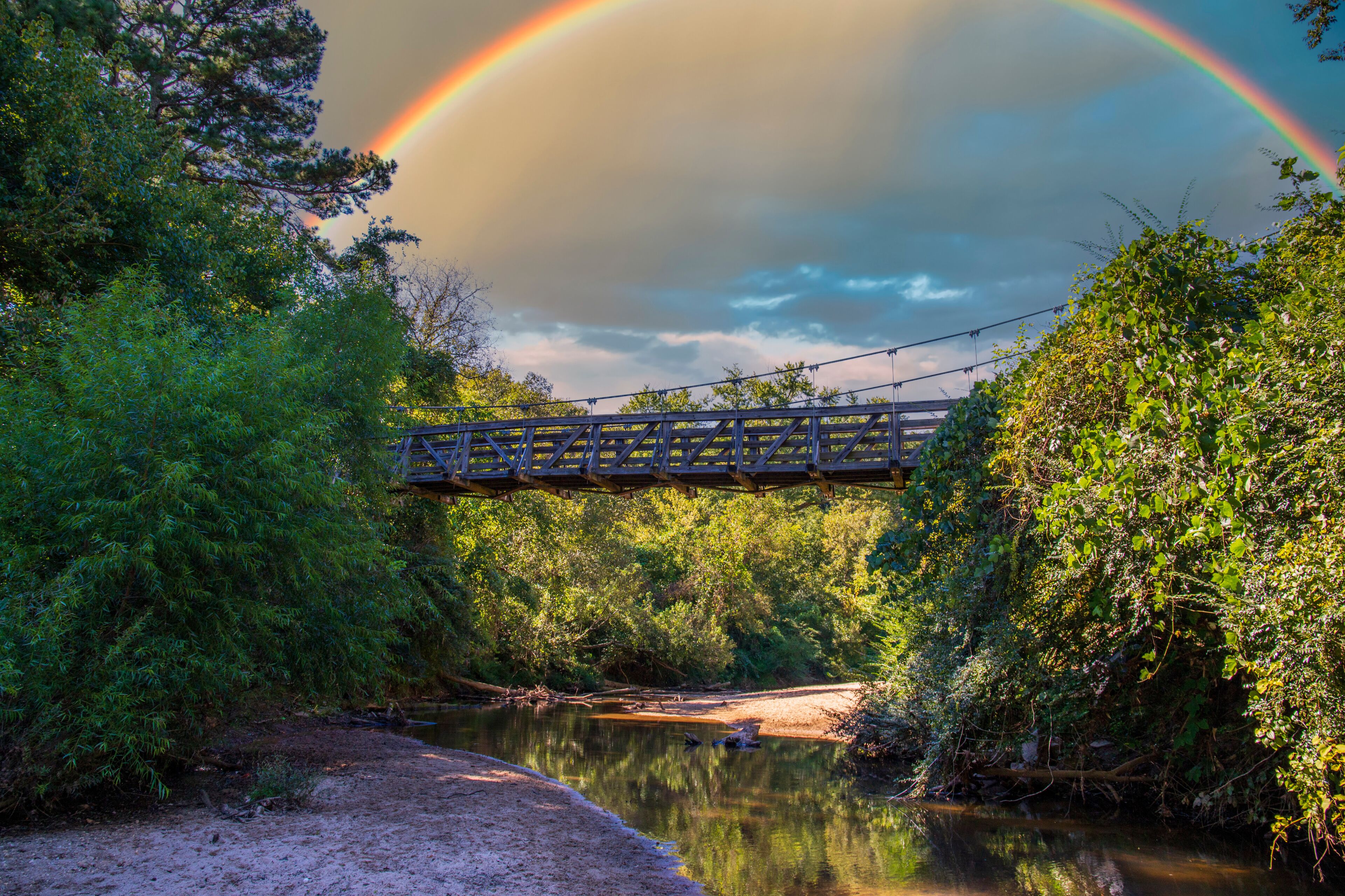 a brown wooden suspension bridge over the clear waters of the South Fork Peachtree Creek surrounded by lush green tree, grass and plants and sand along the banks with blue sky, clouds and a rainbow