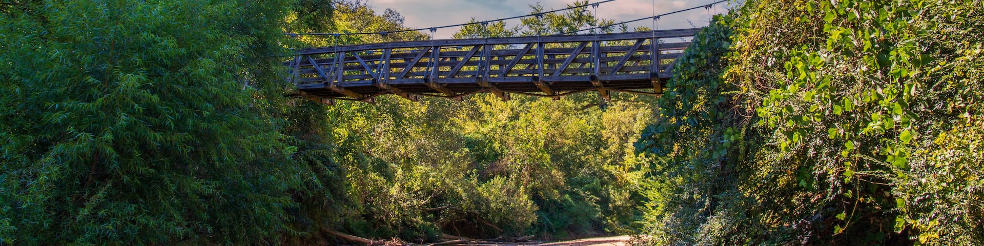 a brown wooden suspension bridge over the clear waters of the South Fork Peachtree Creek surrounded by lush green tree, grass and plants and sand along the banks with blue sky, clouds and a rainbow