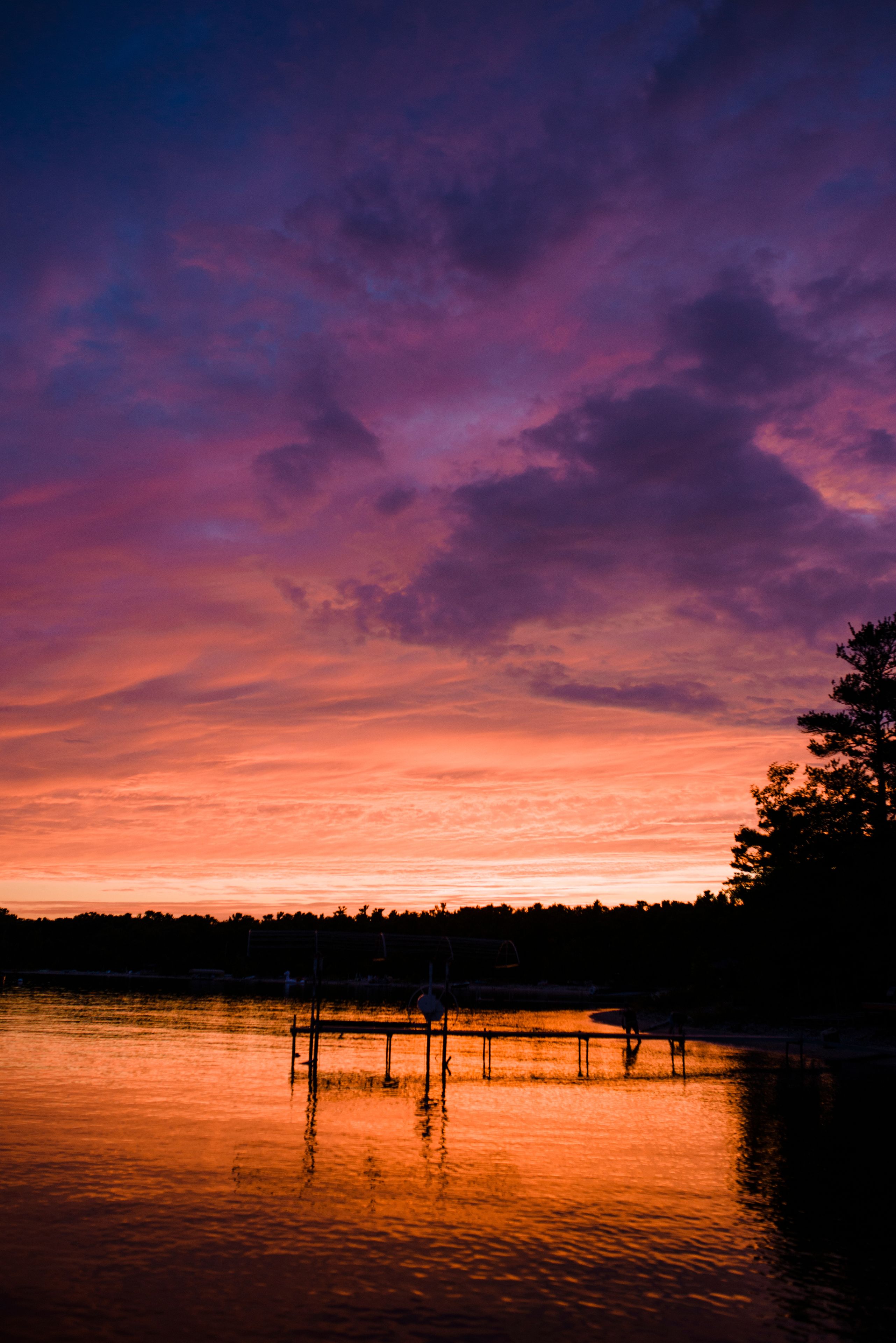 A sunset over a calm lake with a dock and trees silhouetted against the sky.